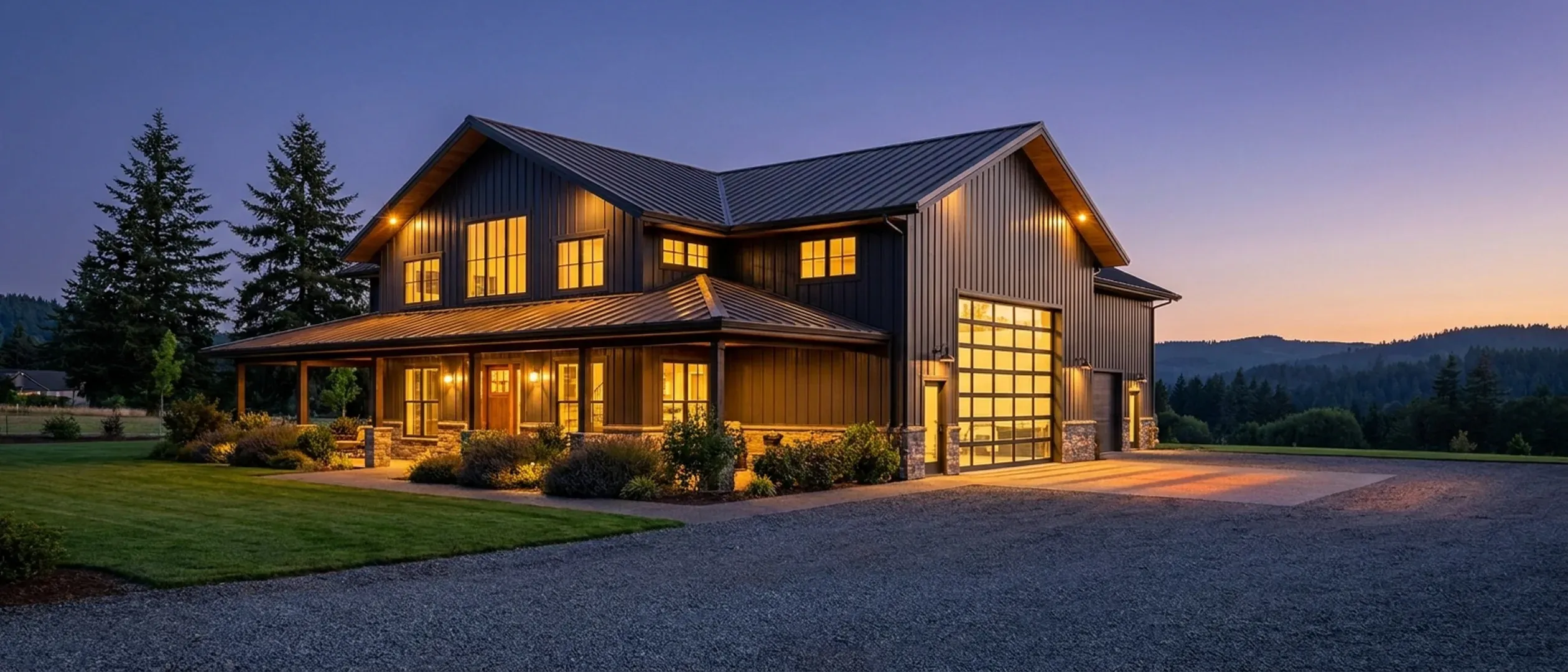 A barndominium at dusk with warm light glowing from every window and exterior lighting along the porch and roofline, tall firs dark against a deep blue twilight sky.