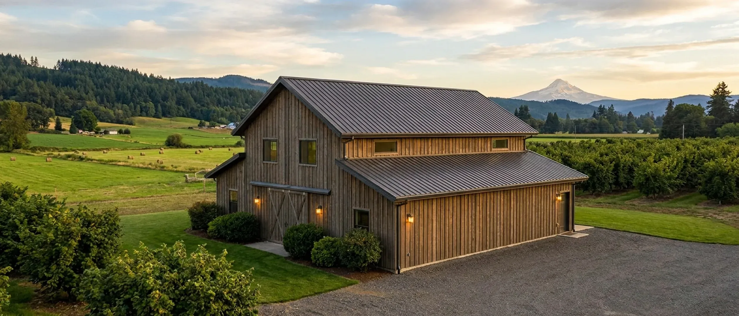 A completed wood-clad barndominium with a standing seam metal roof, gutters and exterior wall lights, green farmland and a snow-capped peak behind.