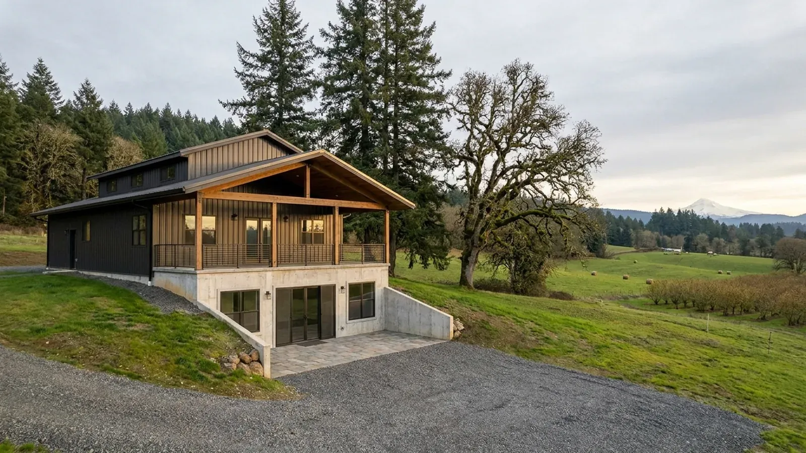 A barndominium with a walkout basement built into a sloping lot, the lower level opening onto a patio at grade, with tall firs behind.