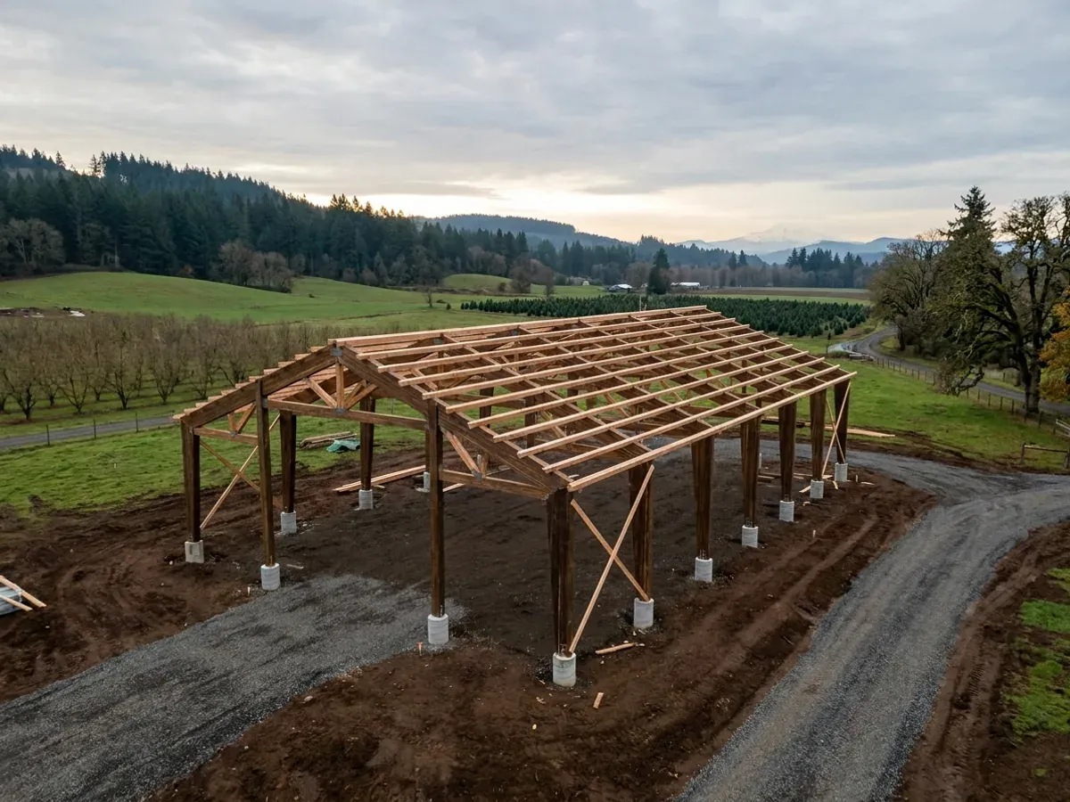 A pole barn home under construction, showing laminated timber columns on concrete piers and wide-span roof trusses with no interior bearing walls.