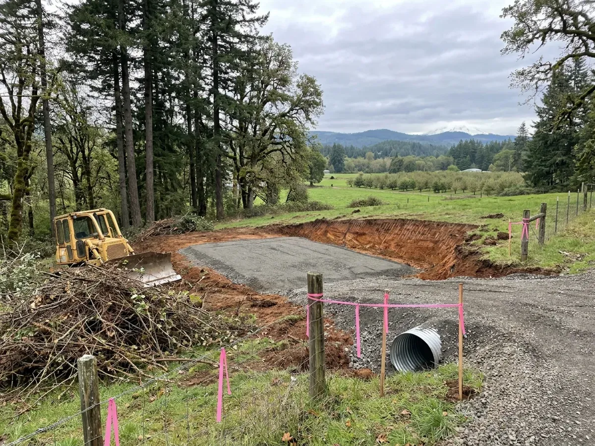 A dozer clearing brush on a rural parcel beside a compacted crushed-rock building pad, with reddish-brown subsoil in the cut and a culvert set at the driveway entrance.